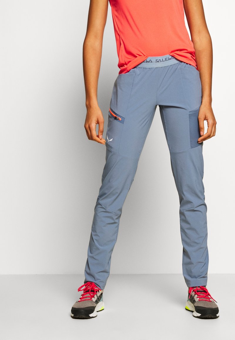 Personne portant un pantalon de randonnée gris avec une poche latérale zippée, un haut corail vif, et des baskets grises avec des lacets roses, debout sur un fond blanc.
