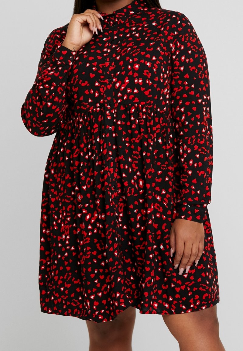 Femme portant une robe noire à manches longues avec des taches en forme de cœur rouges et blanches, boutonnée à l'avant et jupe évasée.