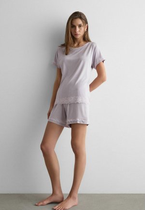 Giovane donna in piedi a piedi nudi che indossa una maglietta a maniche corte lilla chiaro e pantaloncini abbinati con bordi di pizzo davanti a una parete bianca semplice.