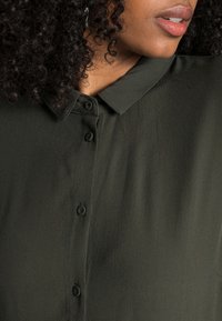 Camisa verde-escura de botão com colarinho pontiagudo, apresentando um tecido suave e liso e seis botões iguais na parte da frente.