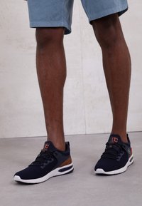 Navy mesh sneakers with brown accents, white outsole, and intricate lacing system. Rounded toe and padded collar for comfort.
