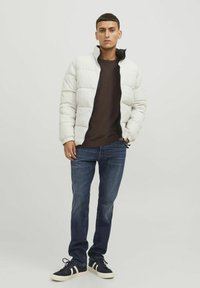 Chaqueta blanca acolchada con cremallera negra, llevada sobre un suéter de cuello redondo marrón. Combinada con jeans azul oscuro y zapatillas negras con rayas blancas.