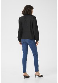 Blouse noire à manches bouffantes, portée avec un jean skinny bleu et des mules à talons noires, fond uni. Accent sur le design des vêtements et la coupe.