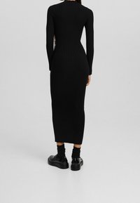 Abito nero a costine con maniche lunghe, silhouette aderente e lunghezza midi. Accentuato da ritagli laterali e abbinato a scarpe nere chunky.