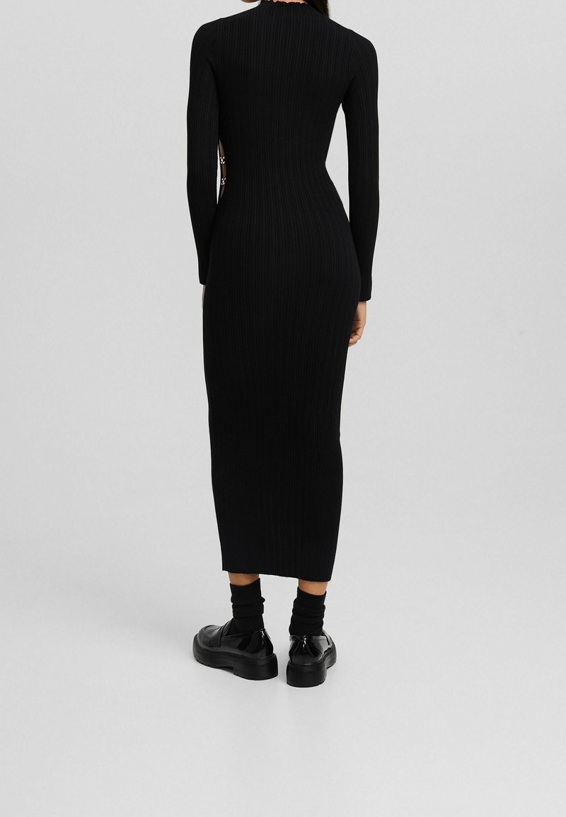 Abito nero a costine con maniche lunghe, silhouette aderente e lunghezza midi. Accentuato da ritagli laterali e abbinato a scarpe nere chunky.
