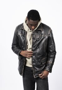 Chaqueta de cuero negra con diseño de doble botonadura, costuras visibles, bolsillos tipo parche y un bolsillo con cremallera, superpuesta sobre una sudadera beige con capucha.