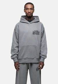 Felpa grigia in misto cotone con tasca frontale, caratterizzata da una stampa grafica sul petto. Abbinata a pantaloni della tuta grigi coordinati.