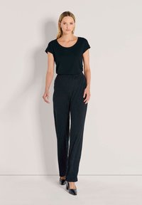 Vrouw in een zwart shirt met korte mouwen, gestructureerde zwarte hoge taille broek en zwarte hakken poserend tegen een effen lichte achtergrond.