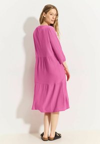Fuchsia Midi-Kleid aus strukturiertem Stoff, mit langen Ärmeln und einem gestuften Design. Kombiniert mit schwarzen Sandalen mit einer Schleifenverzierung.
