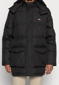 Svart puffjacka med huva, med dragkedjestängning, framfickor med lock och en logotyp-patch på vänster bröst. Slät, isolerad material.