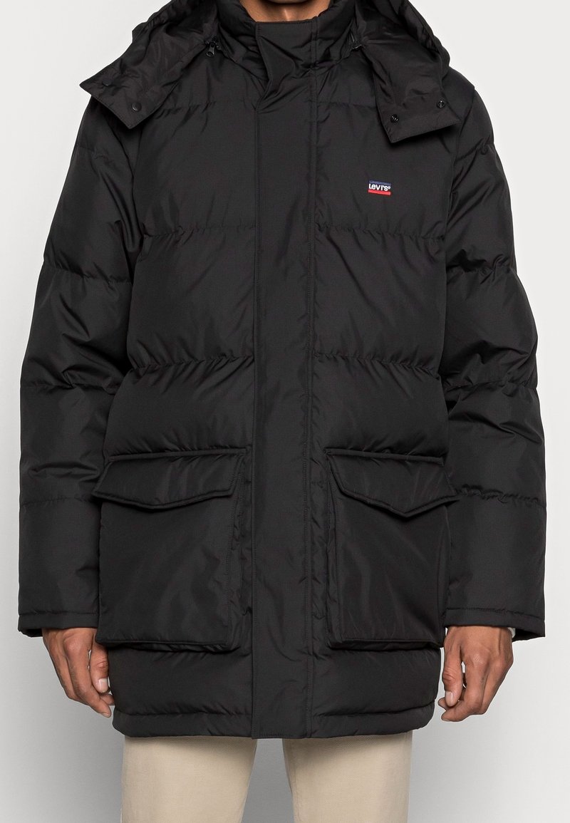 Svart puffjacka med huva, med dragkedjestängning, framfickor med lock och en logotyp-patch på vänster bröst. Slät, isolerad material.