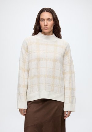 Femme aux cheveux bruns ondulés portant un pull à carreaux blanc et beige et une jupe marron, debout sur un fond blanc uni.