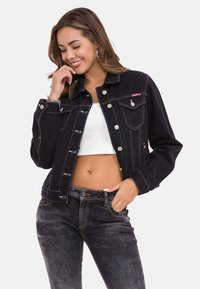 Zwarte denimjacket met contrasterende gele stiksels, knoopsluiting en twee voorzakken. Draagt samen met een witte crop top en zwarte jeans.