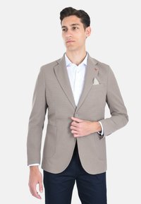 Blazer testurizzato beige chiaro con due tasche frontali, chiusura a un bottone e fazzoletto bianco a contrasto su una camicia bianca.
