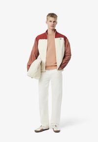 Chaqueta para hombre en dos tonos con una combinación de colores crema y óxido, combinada con una camisa beige clara, pantalones blancos y zapatillas crema. Incluye un bolso.