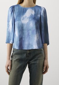 Blouse en soie bleue avec un motif aquarelle, manches trois-quarts et col rond, associée à un jean en denim bleu clair.