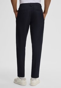Pantaloni blu navy con cintura elasticizzata e due tasche posteriori, caratterizzati da una vestibilità dritta e un tessuto liscio. Indossati con sneakers di colore chiaro.