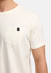 Krämfarbener Baumwoll-T-Shirt mit Rundhalsausschnitt und kurzen Ärmeln. Verfügt über eine linke Brusttasche mit einem schwarzen Etikett. Weiches Material.