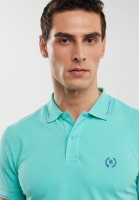 Jasnoturkusowa koszulka polo z teksturowanej tkaniny, z dwoma guzikami i haftowanym logo w ciemnoniebieskim kolorze. Akcentowana cienkimi kontrastującymi liniami kołnierza.