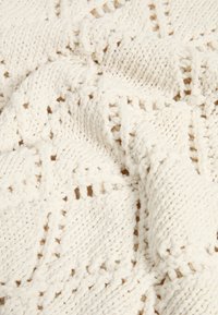 Plaid blanc tricoté présentant un motif texturé avec des mailles ajourées, offrant un aspect doux et confortable. Le matériau semble épais et volumineux.