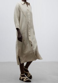 Abito camicia in lino beige con chiusura a bottoni sul davanti, maniche lunghe e leggere fessure laterali. Presenta un dettaglio di bordo ondulato lungo le cuciture.