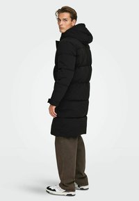 Zwarte pufferjas met een capuchon, voorzien van een gequilte design en een lange snit. Gekleed met donkere broek en zwart-witte sneakers.
