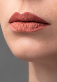 Matte läppfärg i varm brunt ton på släta läppar, som framhäver textur med definierad kontur och subtil naturlig hudton i bakgrunden.