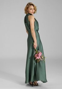 Langes, ärmelloses, grünes Satinkleid mit drapiertem Ausschnitt und offener Rückseite. Das Modell hält einen Strauß mit rosa Blumen und trägt schwarze Absätze.