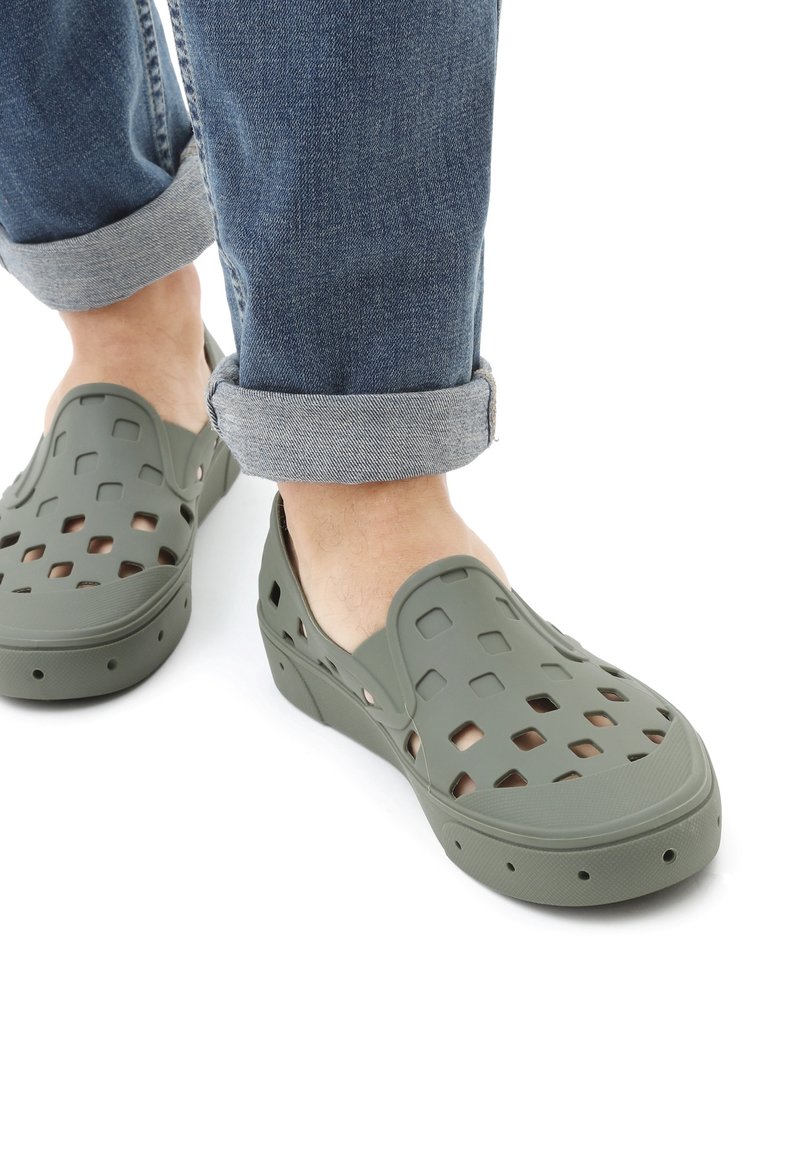 Green slip-on shoes made of rubber with cut-out patterns and a supportive sole. Worn with rolled-up denim jeans.