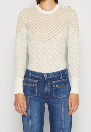 Femme portant un pull en maille blanc et beige à motif chevron et un jean denim bleu avec des accents de chaîne dorée sur les poches avant.