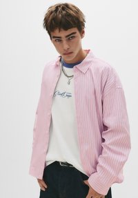 Cămașă cu dungi roz deschis, cu nasturi și mâneci lungi, asociată cu un tricou alb cu text albastru. Modelul poartă blugi negri.