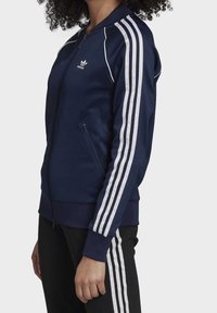 Osoba oblečená v námořnické modré sportovní bundě Adidas s bílými pruhy na rukávech a odpovídajícími černými kalhotami s bílými pruhy na stranách, stojící bokem.