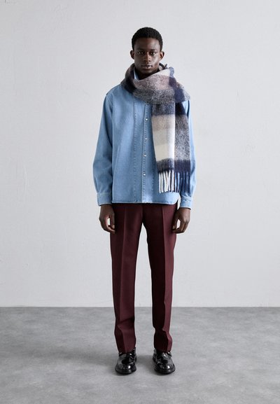 Denim shirt in light blue, red trousers, and a plaid wool scarf in navy, gray, and cream colors; polished black shoes complete the look.