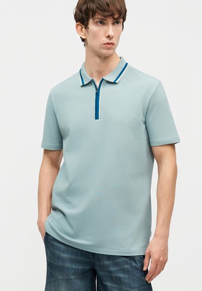 Jeune homme portant un polo à manches courtes bleu clair avec un col rayé bleu et un short en denim foncé, debout les mains dans les poches.