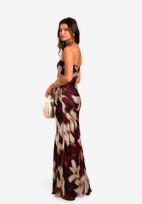 Trägerloses Maxikleid mit floralen Mustern in Nuancen von tiefrot, braun und creme mit glatter Textur; kombiniert mit einer hellen Clutch.