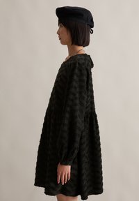 Schwarzes strukturiertes Kleid mit langen Ärmeln und gerafftem Rock, versehen mit einem gewellten Ausschnitt. Getragen mit einer schwarzen Schirmmütze.