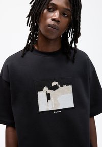 Schwarzes Baumwoll-T-Shirt mit einem strukturierten rechteckigen Aufnäher mit abstraktem beige- und weißem Design sowie einem bestickten Etikett mit der Aufschrift "Eine neue Idee."