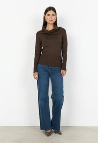 Kvinna som står mot en vit vägg, iklädd en brun cowl neck-tröja, blå vida jeans och zebramönstrade spetsiga skor.