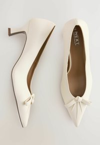 Zapatos de tacón alto blancos con punta afilada y acento de lazo, con parte superior de sintético suave y forro interior marrón.