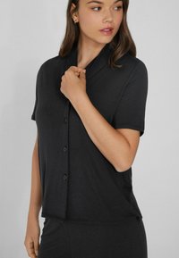 Black short-sleeved button-up top made of soft fabric, featuring a collar and a relaxed fit, with rounded hem and simple black buttons.