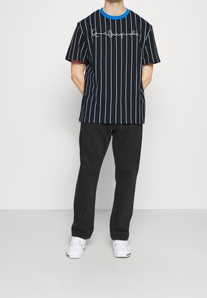 Camiseta de rayas negras con acentos azules, que presenta líneas verticales y un gráfico distintivo en la parte delantera. Se combina con pantalones negros y zapatillas blancas.