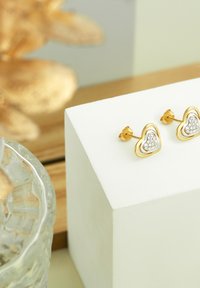 Guld hjerteformede stud øreringe, med en sølvindsats og funklende detaljer, som ligger på en hvid piedestal.