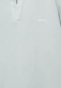 Tissu texturé gris clair avec col en V et petite broderie blanche indiquant "Est. 1921" sur le côté supérieur gauche.