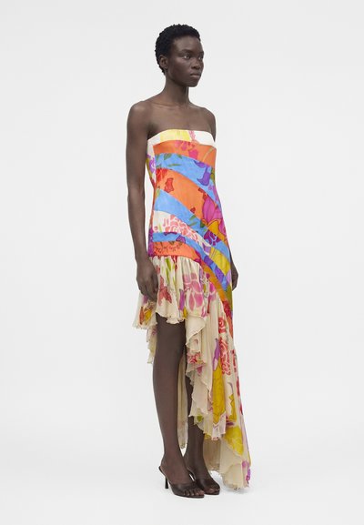 Γυναίκα με ασύμμετρο φόρεμα χωρίς τιράντες με φωτεινά πορτοκαλί, μπλε, ροζ και κίτρινα σχέδια, που φοράει μαύρα σανδάλια με τακούνι.