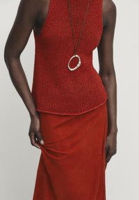 Punainen neulottu hihaton paita, jossa on teksturoitu pinta, yhdistettynä punaiseen mokkihameeseen. Hopeinen soikea riipus roikkuu nahkaketjusta.