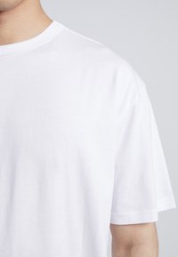 T-shirt bianca in cotone con girocollo, maniche corte e una superficie liscia, mostrata da un'angolazione ravvicinata. Nessun motivo o dettaglio visibile.