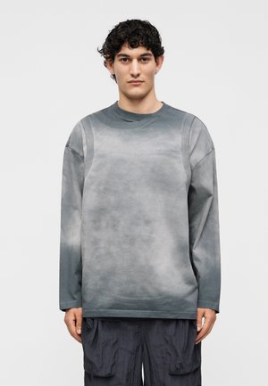 Νεαρός άνδρας με σκούρα σγουρά μαλλιά, φορώντας γκρι μακρυμάνικο πουκάμισο και μαύρο υφασμάτινο παντελόνι, στέκεται μπροστά σε ένα απλό φωτεινό φόντο.