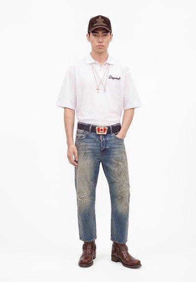 Homme debout portant un polo blanc, un jean bleu délavé, des bottes marron, une casquette marron, deux colliers croix et une ceinture avec une boucle rouge et blanche.