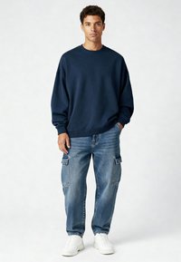 Tumši zila pūkainā džemperis ar ribotām piedurknēm un apakšmalu, savienots ar gaiši ziliem kargo džinsiem ar sānu kabatām un brīvu piegriezumu. Baltas sporta apavi.