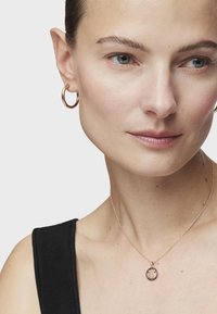 Pendiente de aro en oro rosa y collar delicado con cadena que presenta un colgante circular con un diseño texturizado en tonos oscuros y claros. Top negro.